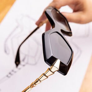 Hands holding the finished Serpenti Viper black acetate sunglasses with metallic temples over a sketch.